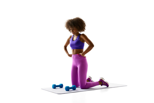 穿着紫色运动服的女人跪在蓝色哑铃旁边的垫子上，俯视着白色的工作室背景。图片下载