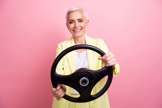 照片中成熟的女商人穿着黄色的正装优雅的驾驶握方向盘慢动作孤立在粉红色的背景图片下载