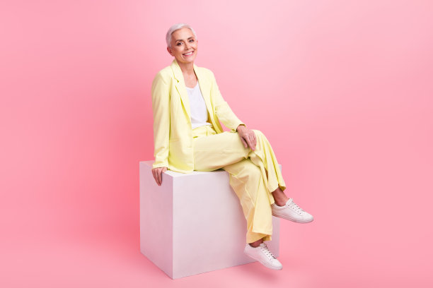 全尺寸照片的时尚积极愉快的女人穿着黄色西装坐在白色立方体交叉腿孤立在粉红色的背景图片下载