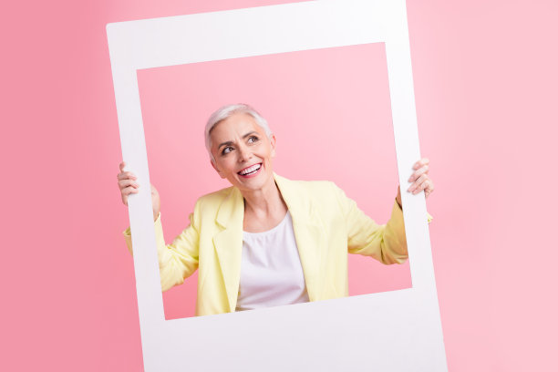 白色发型的漂亮女人的照片，穿着黄色的西装，拿着相框看，在粉红色的背景上提供了空白的空间图片下载