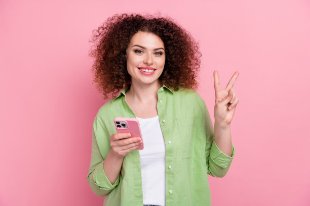 照片中，有趣可爱的女人烫发，穿着时尚的衬衫，拿着智能手机，在粉红色的背景上显示v字图片下载