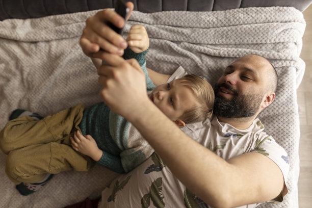 爸爸和小儿子笑着看着手机上的照片。他们躺在卧室的床上拍照。父亲拥抱儿子。他们做鬼脸。一个深色头发和浓密胡须的男人和一个浅色头发和眼睛的男孩图片下载