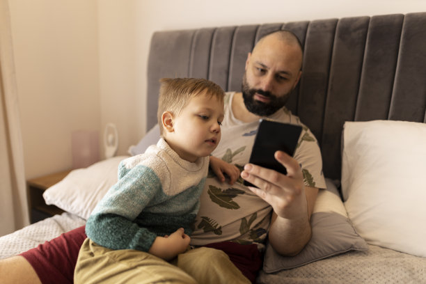 爸爸和小儿子笑着看着手机上的照片。他们坐在卧室的床上拍照。父亲拥抱儿子。他们做鬼脸。一个黑发浓密的男人和一个金发碧眼的男孩。焊接时间图片下载