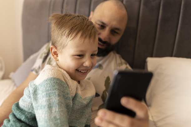 爸爸和小儿子笑着看着手机上的照片。他们坐在卧室的床上拍照。父亲拥抱儿子。他们做鬼脸。一个黑发浓密的男人和一个金发碧眼的男孩。焊接时间图片下载