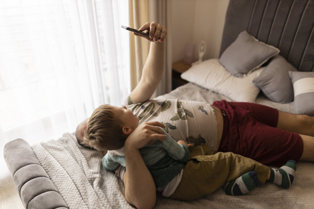 爸爸和小儿子笑着看着手机上的照片。他们躺在卧室的床上拍照。父亲拥抱儿子。他们做鬼脸。一个深色头发和浓密胡须的男人和一个浅色头发和眼睛的男孩图片下载