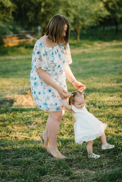 母亲牵着孩子的手，转着圈。夕阳下，妈妈和小女孩在公园里玩耍。妈妈和孩子花时间在一起。初学走路的女儿在暑假的田野里拥抱母亲的照片图片下载