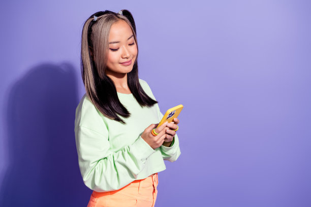 照片中，中国年轻快乐的女学生穿着绿色运动衫，在紫色背景下独自浏览智能手机发短信图片下载