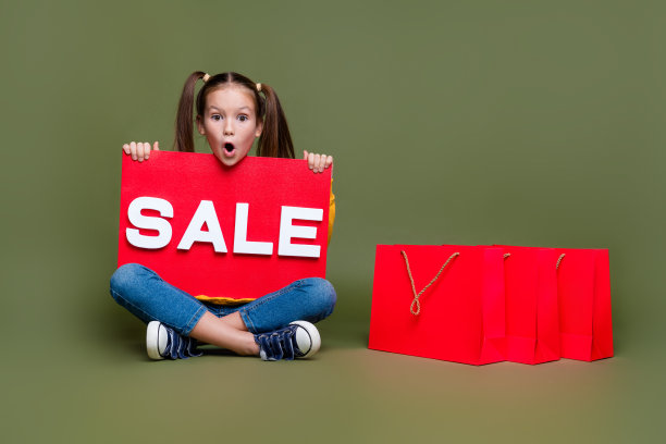 全长照片无言的孩子马尾辫发型坐在地板上持有销售海报盯着销售孤立的卡其色背景图片下载