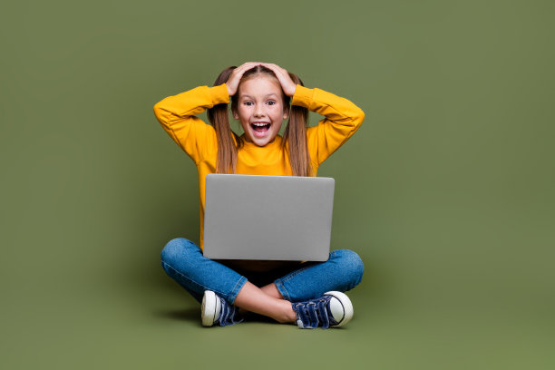 印象深刻的马尾辫头发的孩子的全长照片坐在地板上的腿上的笔记本电脑，手臂在卡其色背景孤立的头图片下载