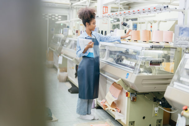 美国女设计师在纺织厂与几台织布机一起工作的全身照片，编织线，拿着一张笔记清单，检查计算机控制的线轴。图片下载