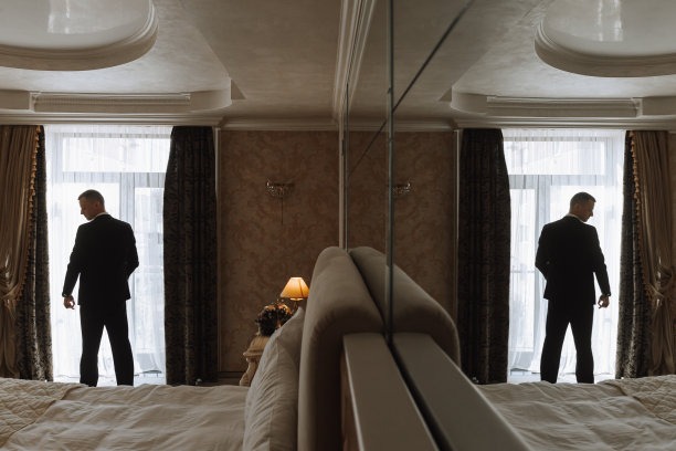 年轻人在他的房间里，在酒店房间的窗户附近，年轻的商人在背光，反射在镜子里。新郎正在为婚礼做准备图片下载