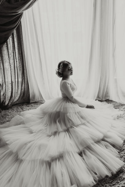 酒店房间里，一位美丽的新娘身穿白色婚纱，拖着长长的裙摆。新娘靠着一扇大窗户坐在地板上。黑白照片。图片下载