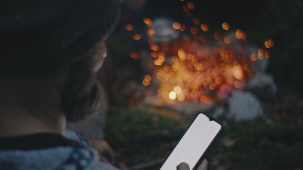 假期在森林露营时，一名男子在营火旁使用智能手机图片下载