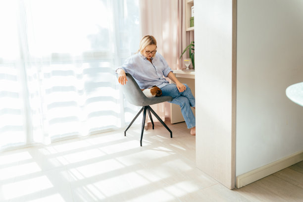 一名女子穿着衬衫，光着脚，站在一所房子的窗户旁，手里拿着一台笔记本电脑。气氛轻松，旁边还有一只狗。女性在家网上购物图片下载