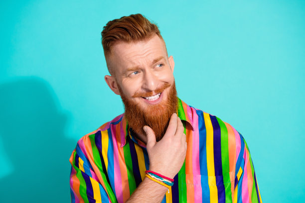 照片中有吸引力的积极的男人穿着时髦明亮的衣服，手臂触摸长长的红胡子，在青色的背景上看起来空无一物图片下载