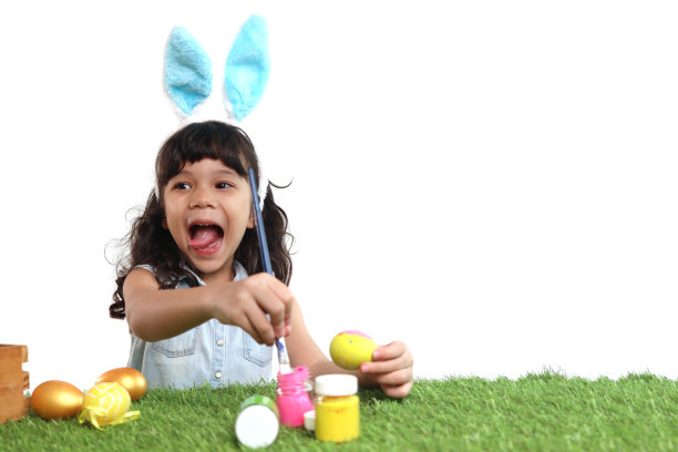 可爱的小女孩长着蓝色的兔子耳朵，用刷子在绿色的草地上画鸡蛋，快乐的孩子在白色的背景上庆祝复活节。快乐的孩子在复活节庆祝活动中玩得很开心。图片下载
