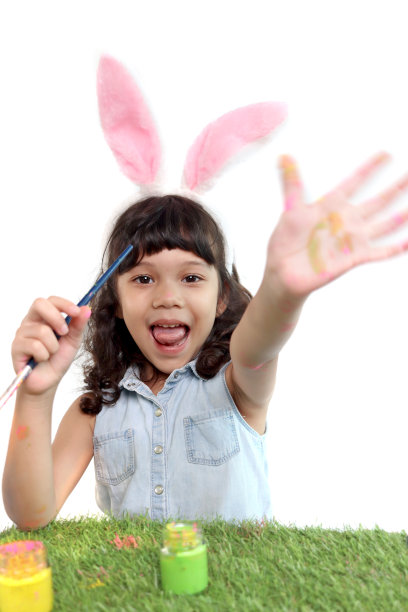 可爱的小女孩有粉红色的兔子耳朵，用刷子在绿色的草地上画鸡蛋。快乐的孩子在白色背景上庆祝复活节。快乐的孩子在复活节庆祝活动中玩得很开心。图片下载