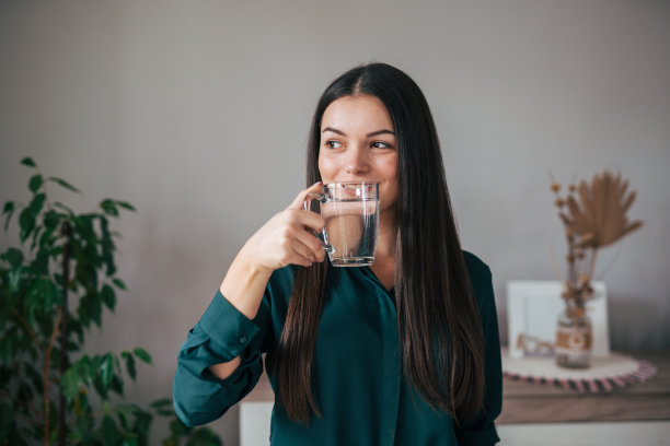 一张年轻女子站在客厅用玻璃杯喝水的照片。图片下载
