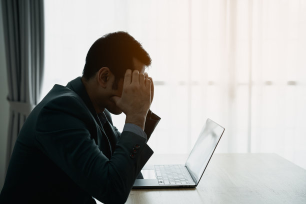 剪影悲伤抑郁严重的亚洲商人有压力的抑郁症悲伤的时间在办公室桌子上的笔记本电脑工作。他看着屏幕，脸上带着沮丧的表情。图片下载