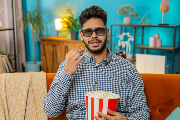 印度男子戴着3D眼镜，吃着爆米花，坐在家里的沙发上看电影图片下载