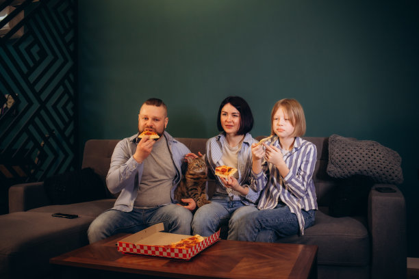 一家人在家里共度美好时光，看起来既开心又兴奋。妈妈，爸爸和女儿玩得很开心，吃披萨，看电视上的体育锦标赛。相聚，家的舒适，爱的概念。图片下载