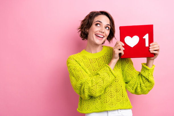 女孩穿着黄色套头衫的照片，手持红色方块心订阅符号分享内容，在粉红色背景上看起来空白图片下载