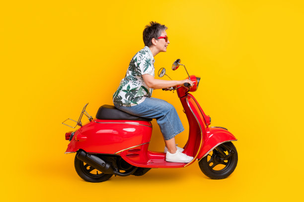 漂亮的退休女士的全身照片驾驶快速红色摩托车穿着时尚的热带印花服装孤立的黄色背景图片下载