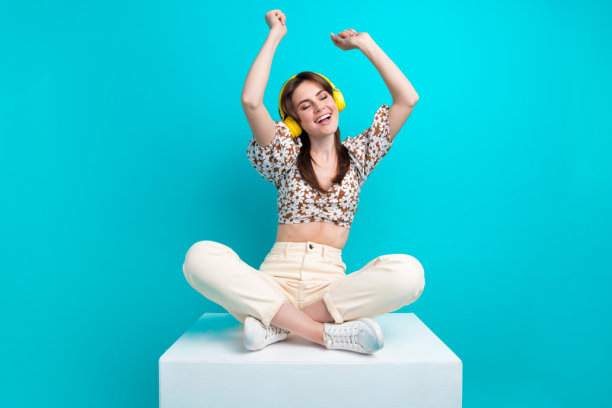 照片中闪亮可爱的女人戴着印花顶，坐在讲台上听歌，耳机孤立的蓝绿色背景图片下载