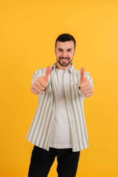 垂直的照片。在黄色背景的摄影棚里，年轻人对着相机竖起大拇指，微笑着。男性摆姿势，用双手微笑着做一个ok的手势，看起来很自信图片下载