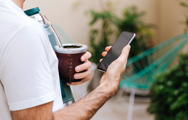 一个拿着葫芦和保温瓶的男人手里拿着一部手机。传统的南美热饮。图片下载