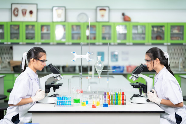 在实验室工作的两名亚洲女科学家，用显微镜观察细胞以生产疫苗，他们面对面坐在一张桌子上，桌子上有测试设备、玻璃管、烧杯等。图片下载