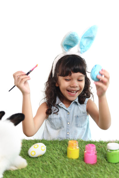可爱的小女孩长着蓝色的兔子耳朵，用刷子在绿色的草地上画鸡蛋，快乐的孩子在白色的背景上庆祝复活节。快乐的孩子在复活节庆祝活动中玩得很开心。图片下载