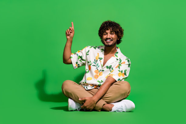 英俊的年轻人的全身照片坐在地板点上穿时髦的柑橘印花服装孤立的绿色背景图片下载