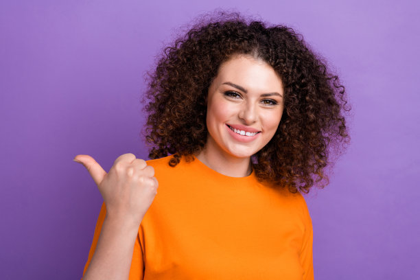 照片中心情好可爱的女子穿着橙色的t恤，露出拇指空白的空间孤立的紫色背景图片下载
