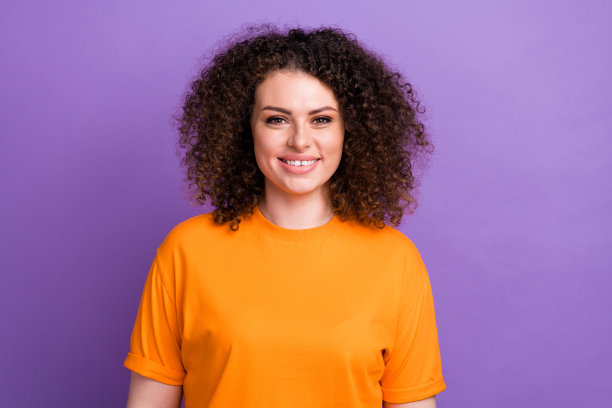 照片中心情积极的女人穿着橙色t恤微笑，露出白色的牙齿孤立的紫色背景图片下载