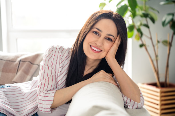 漂亮开朗的照片无忧无虑的女孩灿烂的微笑坐在沙发上，手摸脸颊阳光窗户公寓里面图片下载