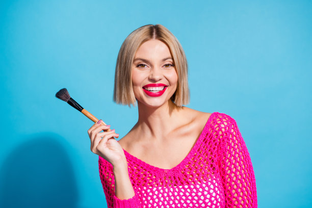 漂亮的年轻女孩的照片肖像拿着画笔化妆师穿着时尚的粉红色针织服装孤立的蓝色背景图片下载