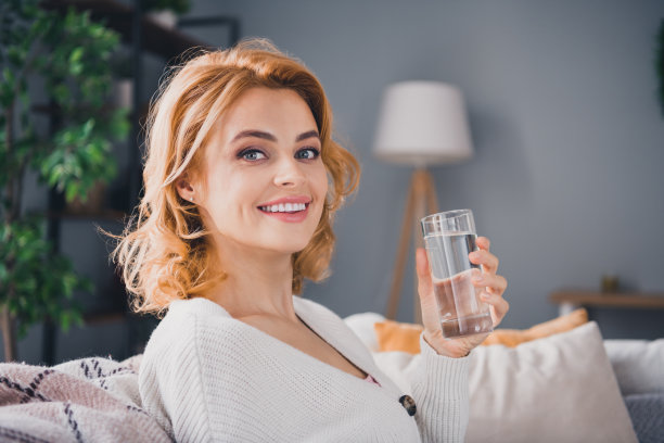 可爱的积极的女孩的资料照片坐在沙发上灿烂的微笑拿着玻璃杯水度过在室内公寓消遣图片下载