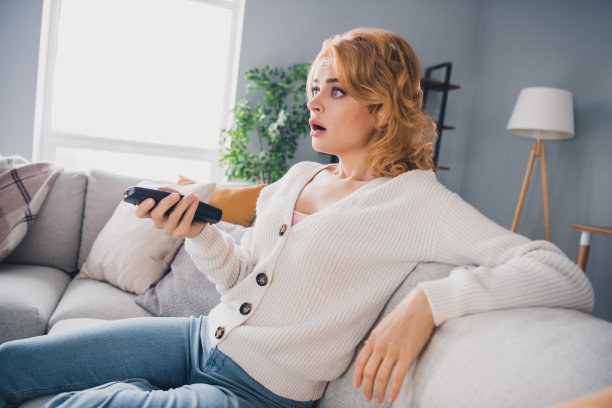 印象深刻的哑口无言的女孩的侧面照片坐在沙发上拿着遥控器张开嘴看电视明亮的房子里图片下载