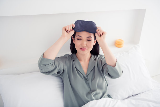 漂亮的年轻女孩躺在床上的照片肖像，摘下面具，穿着时髦的灰色睡衣，孤立在舒适的现代白色卧室里图片下载