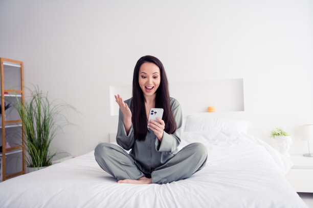 照片肖像迷人的年轻女子坐在柔软的床上抱着装置惊奇地穿着时髦的灰色睡衣隔离在白色现代室内设计图片下载