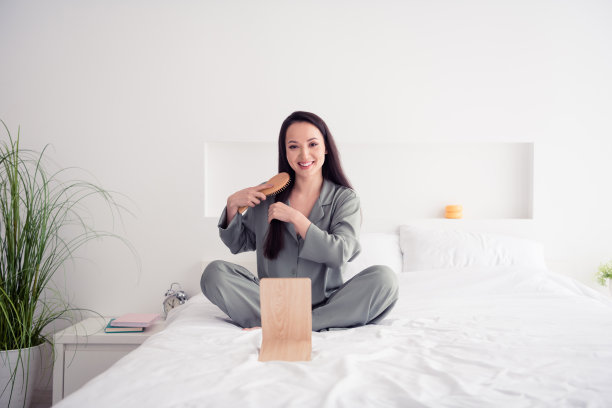 照片肖像迷人的年轻女子坐在柔软的床上刷头发镜子穿时髦的灰色睡衣隔离在白色的现代室内设计图片下载