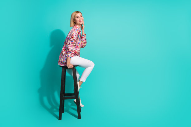 年轻女子的全尺寸照片，坐在椅子上触摸皮肤空的空间穿着时尚的粉红色衣服孤立的海蓝宝石色背景图片下载
