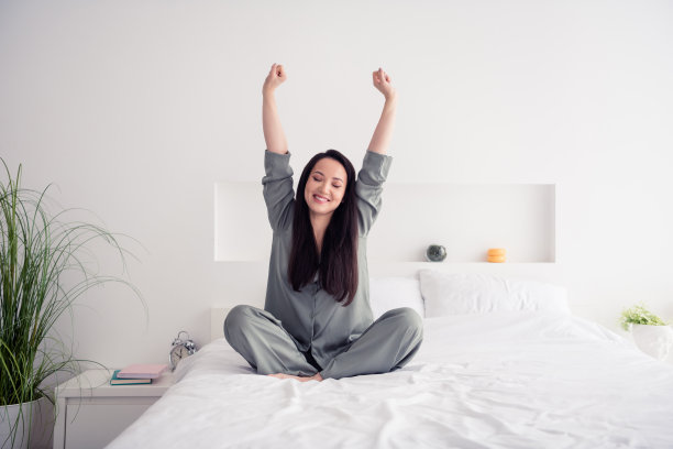 可爱的年轻女士的照片肖像睡前伸展手臂坐在床上穿时髦的灰色睡衣隔离在白色的卧室室内图片下载