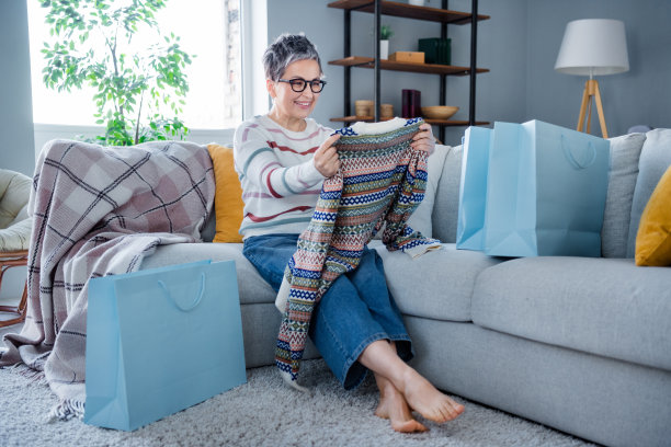 兴奋积极的老太太的全尺寸肖像坐在沙发上打开新毛衣店订购袋现代室内房子里面图片下载