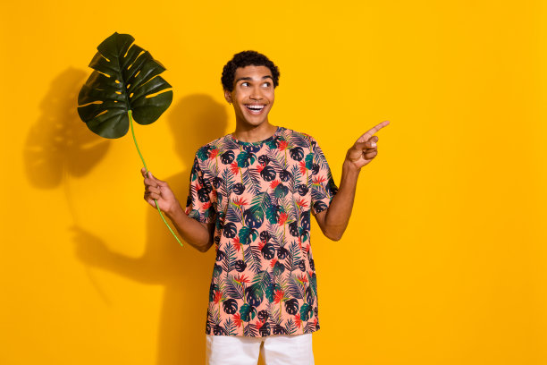 照片中的年轻人用手指指着空旷的空间，拿着热带的绿叶，展示了在黄色背景上孤立的野生树木森林图片下载
