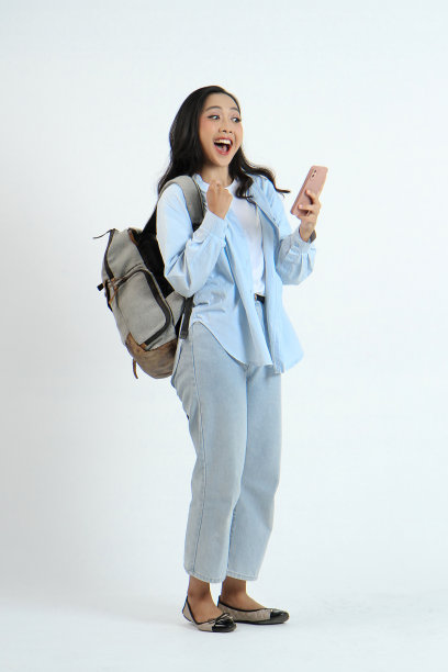 这是一个快乐的亚洲印尼女人的全裸照片，她穿着休闲的衣服，拿着智能手机和背包，背景是孤立的图片下载