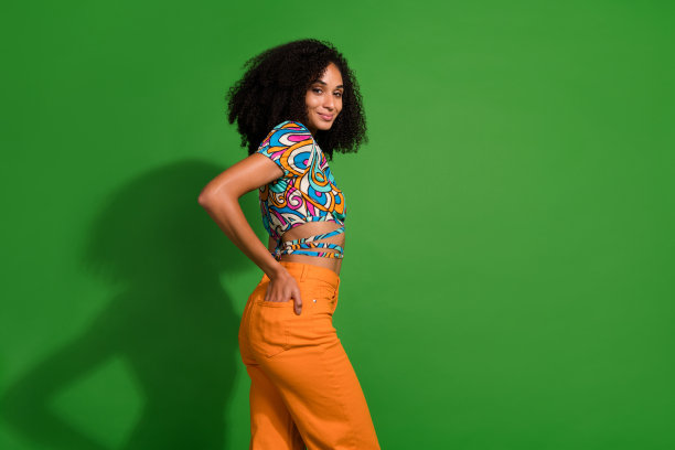 年轻漂亮的女孩的照片，波浪的头发，手臂进入橙色的裤子，摆姿势像一个品牌的模特服装孤立的绿色背景图片下载