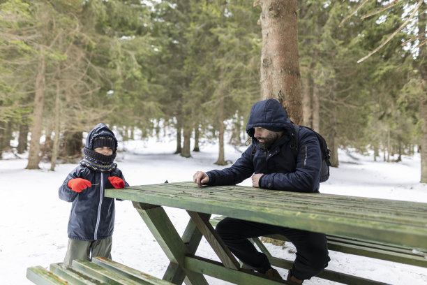 一个快乐的男孩和他的爸爸在森林里玩。他穿着一件深色夹克，戴着红色手套。他们坐在野餐桌上。冬天,下雪了。库存图片图片下载