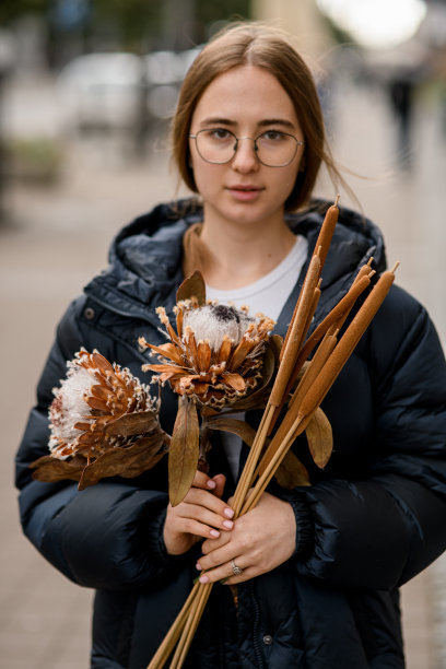 一个棕色头发，戴着眼镜的美丽女孩的照片，她站在城市街道上，手里拿着一束花图片下载